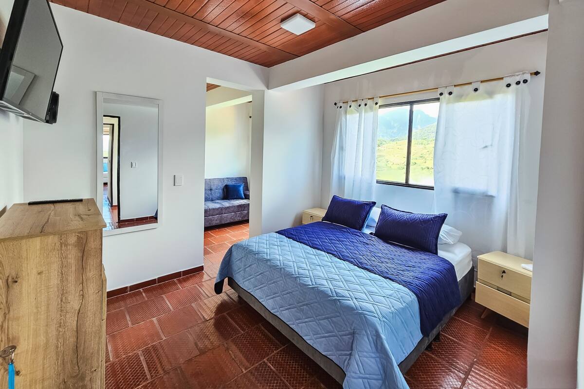 A bedroom with a wooden ceiling, a bed with blue and white bedding, a nightstand, a TV mounted on the wall, and a window with a view of greenery.