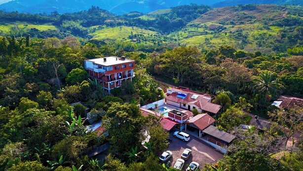 Una casa de varios pisos con piscina y cochera, rodeada de abundante vegetación y colinas.