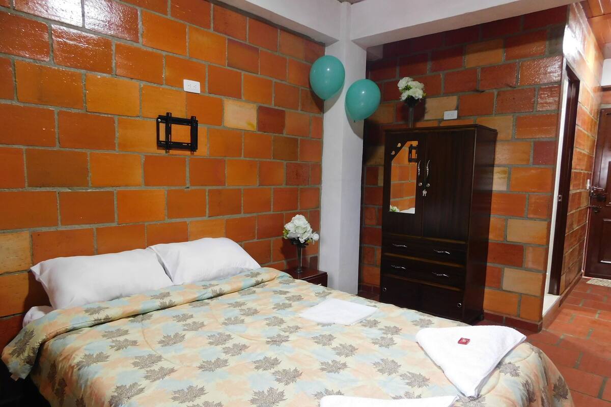 A room with a brick-patterned wall, a bed with a patterned bedspread, a wooden wardrobe, and two balloons.