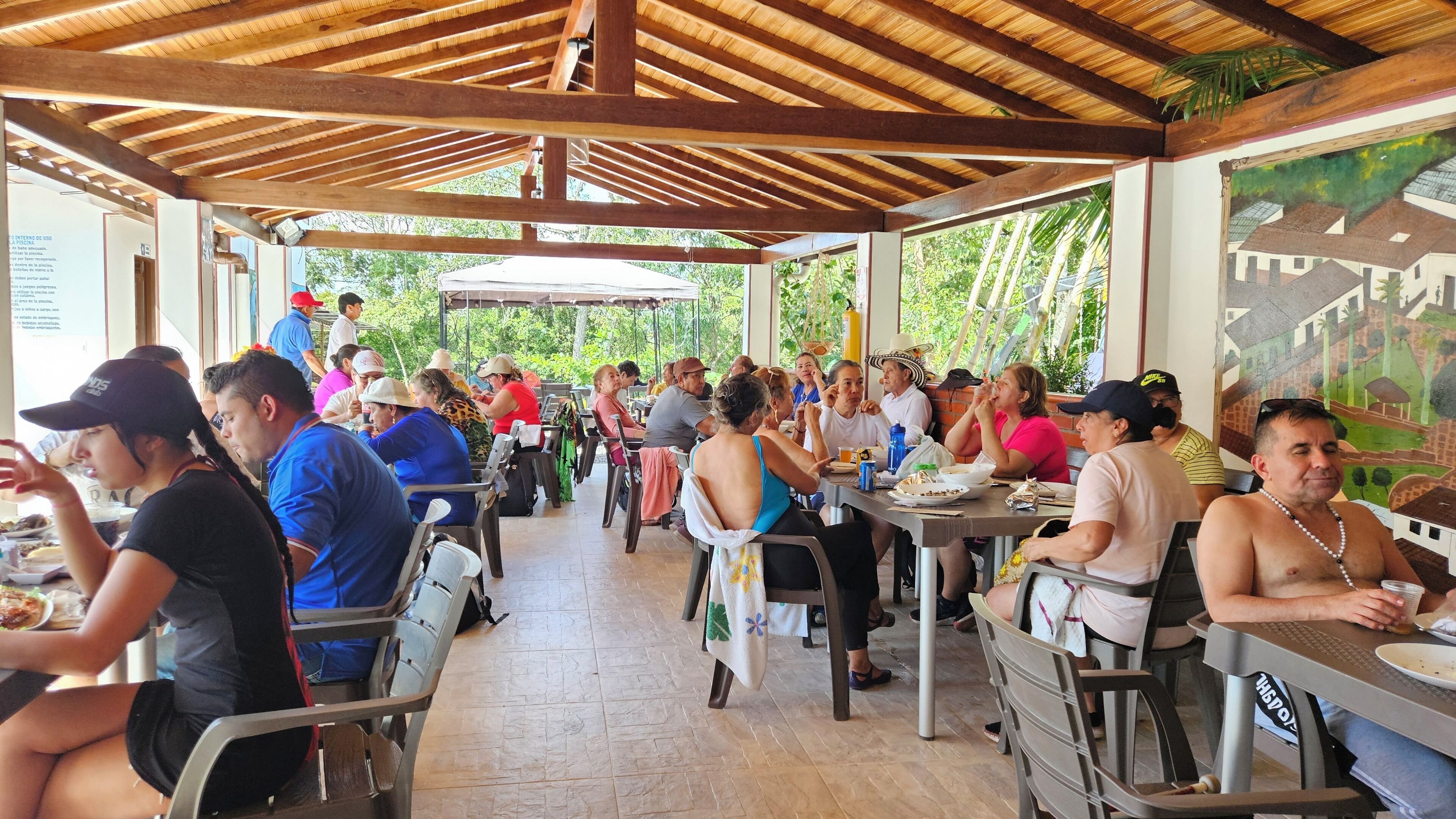 Un espacio de comedor al aire libre cubierto, con mesas y sillas, rodeado de follaje.