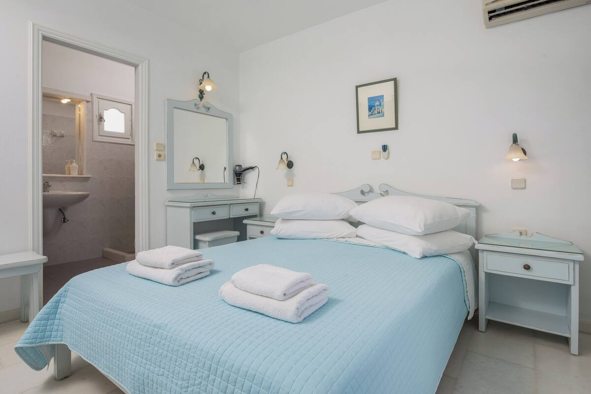 Un dormitorio con una cama, mesitas de noche blancas, una mesilla auxiliar, un espejo y un cuadro colgado en la pared.