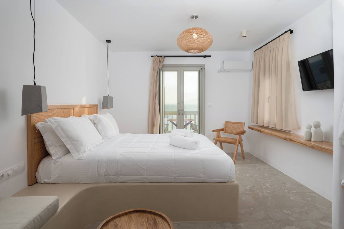 Un dormitorio moderno con una cama grande, cabecera de madera, una mesita de madera, una silla, televisor montado en la pared y un balcón con vistas.