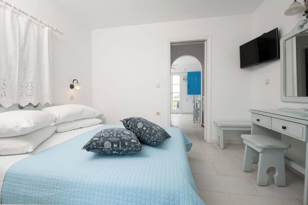 Un dormitorio con cama, almohadas, una mesita de noche, un banco, un televisor y un espejo.