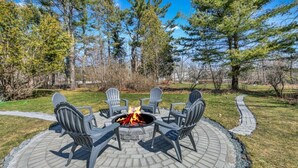 Un espacio circular para sentarse al aire libre con un brasero rodeado de sillas Adirondack, situado en un patio empedrado con vistas a una zona boscosa.