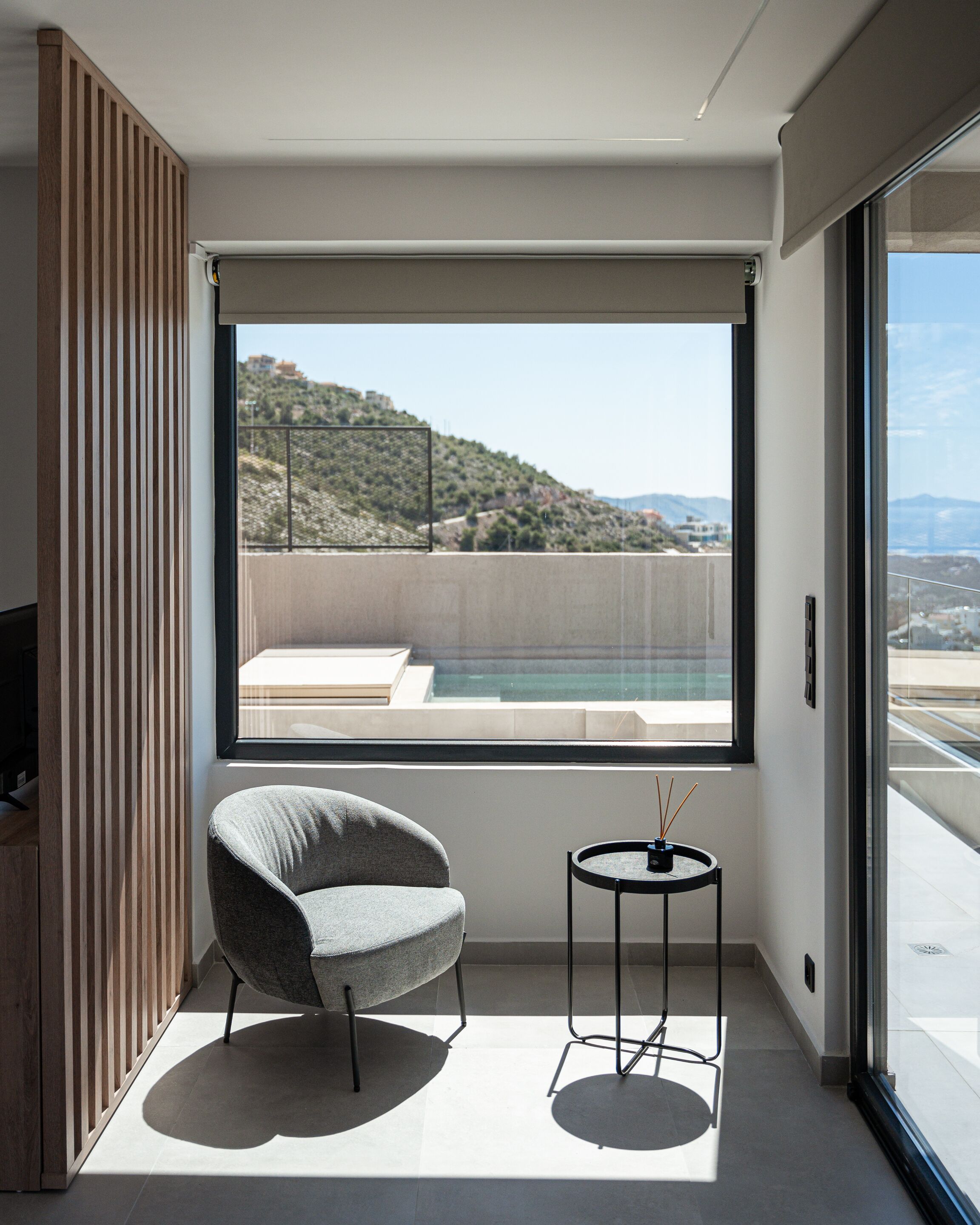 A modern room with a large window, a grey armchair, a small round table, and a wooden slat wall.