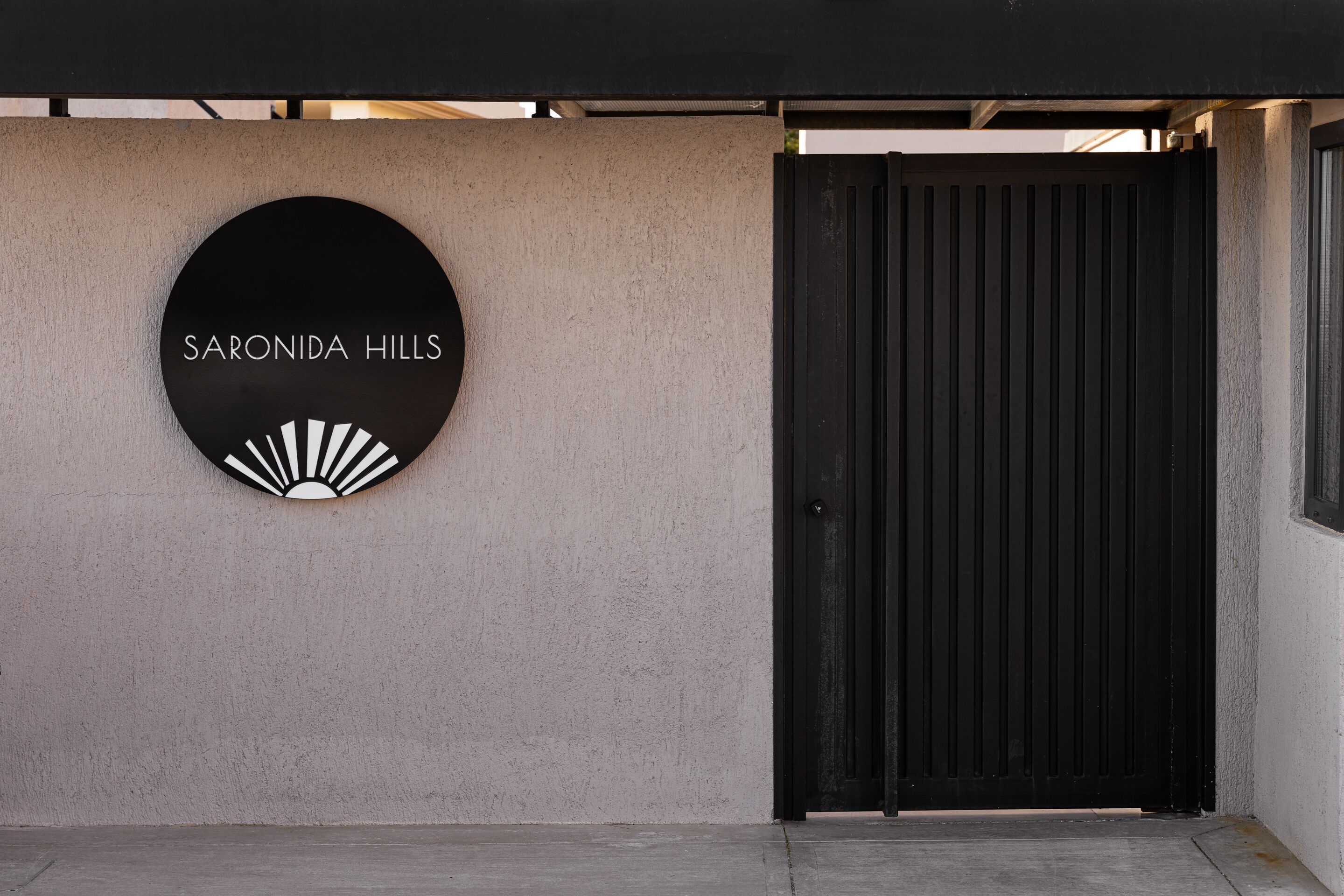 Ein schwarzes, kreisförmiges Schild mit der Aufschrift 'SARONIDA HILLS' und einem Sonnendesign, an einer Betonwand neben einer schwarzen Tür angebracht.