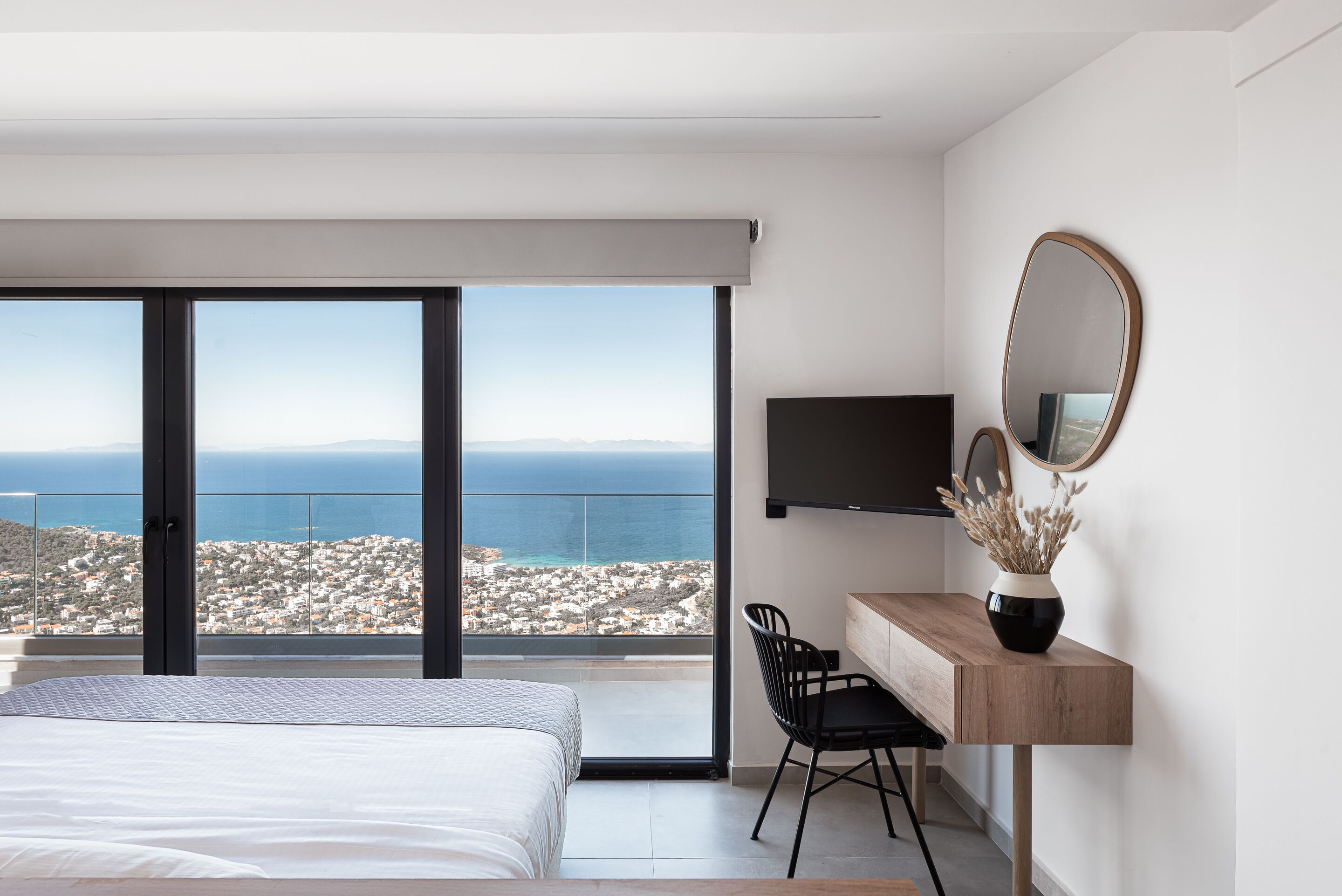 A bedroom with a large bed, a desk with a chair, a TV, and a view of the city and sea through a balcony.
