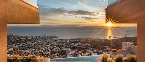 Un bâtiment moderne doté d’une piscine à débordement sur le toit, offrant une vue sur une ville côtière au coucher du soleil.