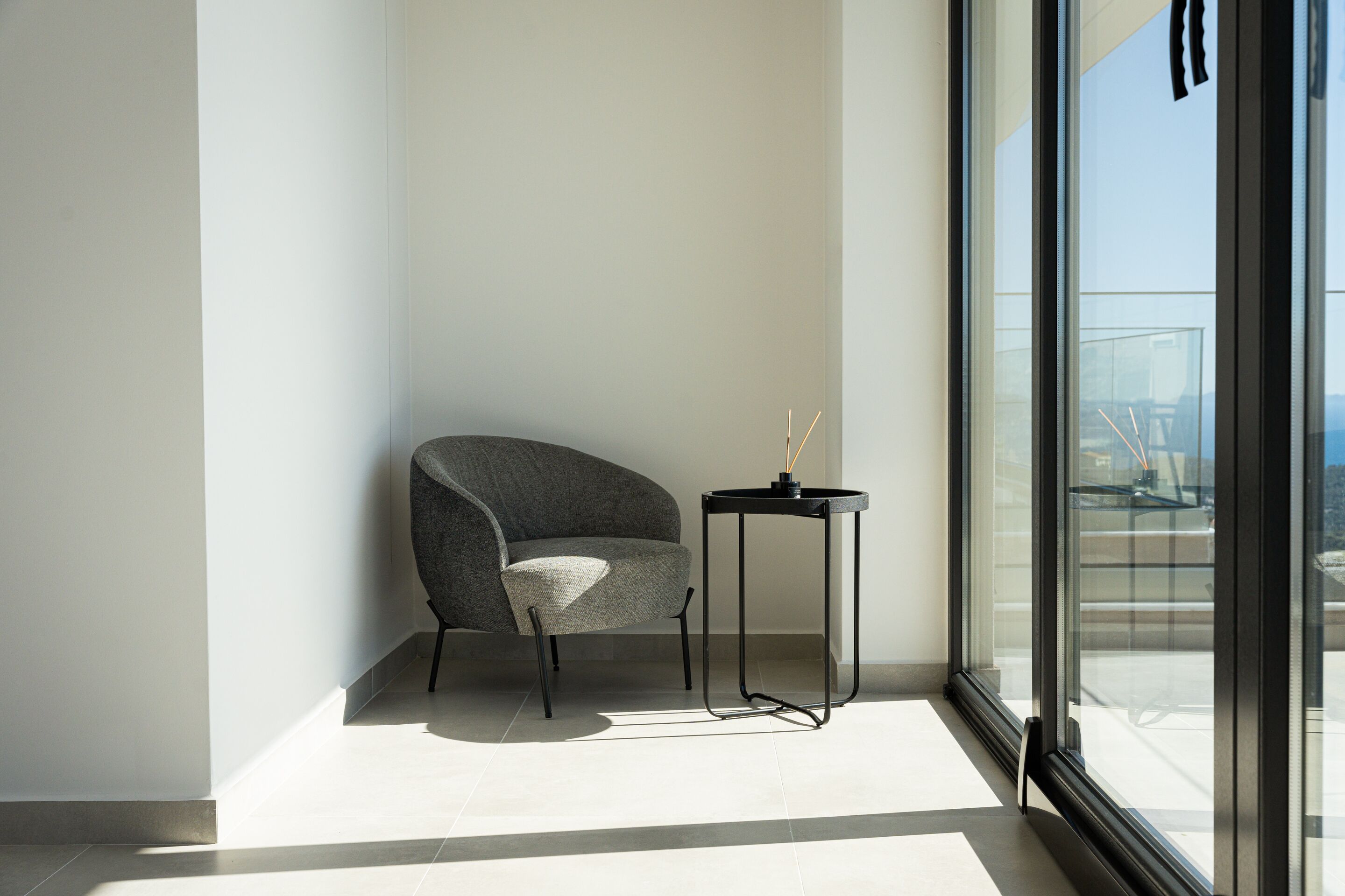 A modern room with a curved grey chair, a small black side table, and a large window offering a view of the city.