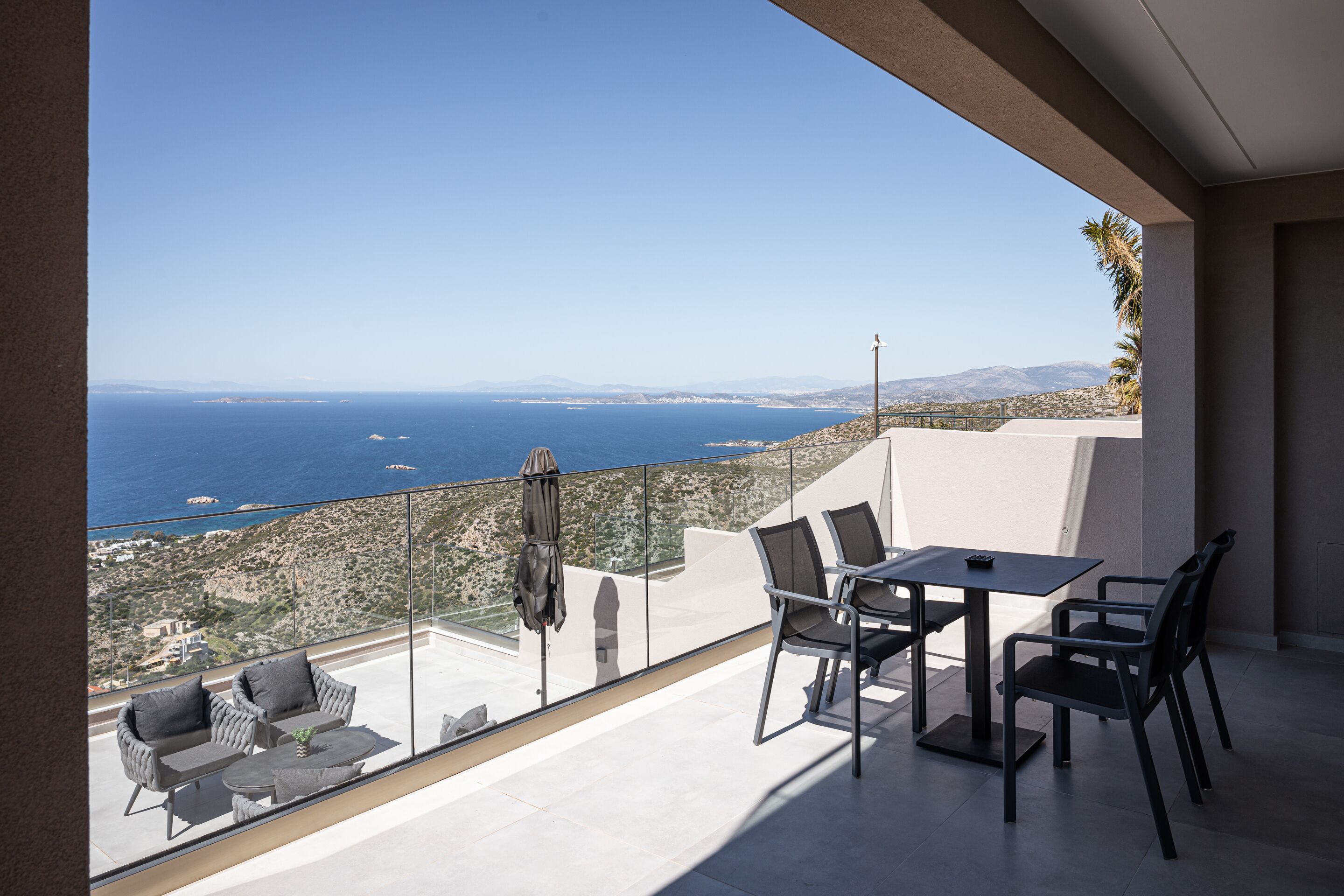 A balcony with a table and chairs, overlooking a coastal landscape with mountains and the sea.