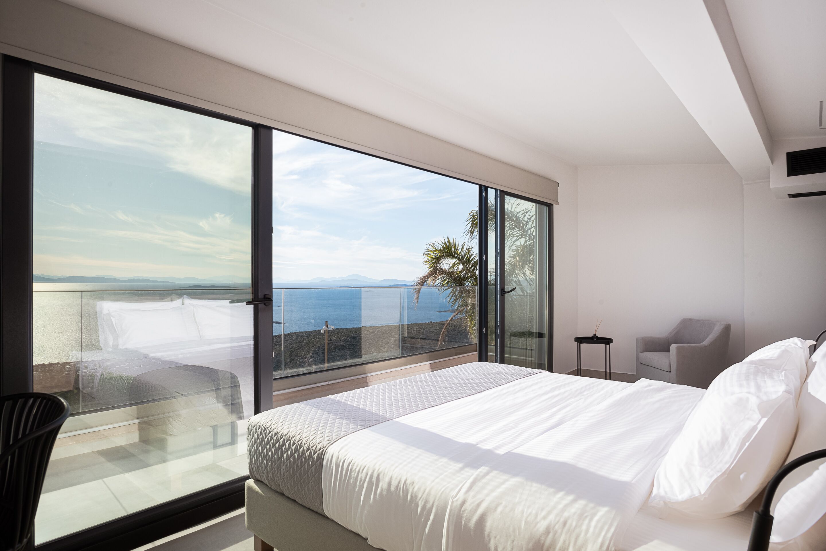 A modern bedroom with a large bed, a glass balcony, and a view of the sea.