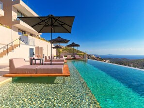 A luxurious infinity pool with clear water, outdoor seating, and umbrellas, overlooking a scenic hillside. - Saronida Hills (Saronikos)