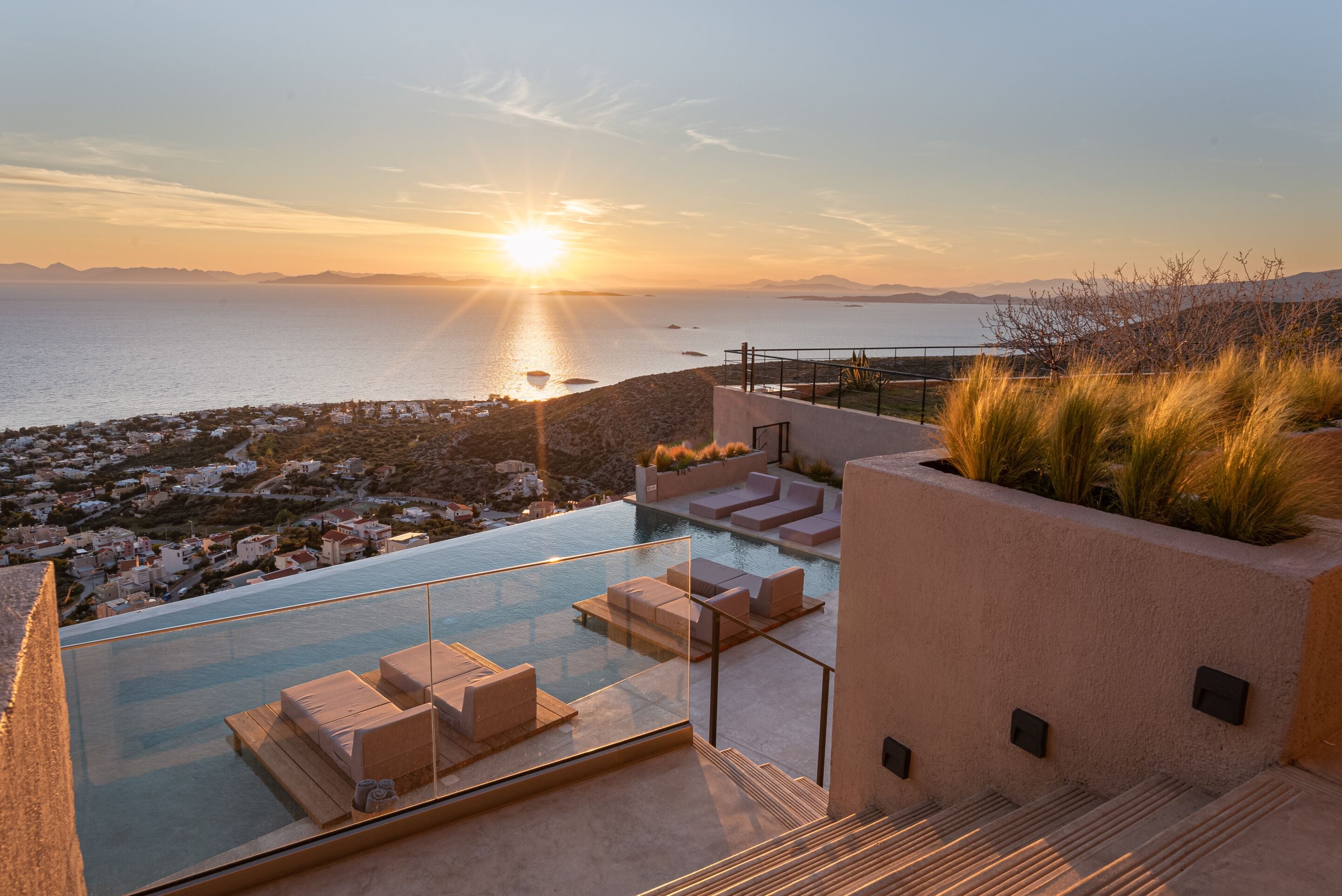 Ein moderner Infinity-Pool mit Glasrand, der auf eine Küstenstadt bei Sonnenuntergang blickt.