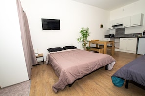 Une petite pièce comprenant un lit, une petite table, une chaise, une plante et un coin cuisine.