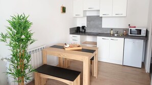 Eine kleine Küche mit weißen Schränken, einer Mikrowelle und einer Kaffeemaschine. Ein kleiner Topfpflanze und ein Holztisch mit einer Bank stehen dort.