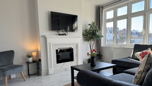 A modern living room with a flat-screen TV mounted above a fireplace, a grey sofa, a coffee table, and a potted plant.