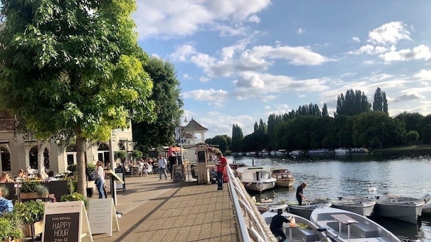 A riverside restaurant with outdoor seating, boats docked nearby, and a sign indicating happy hour.