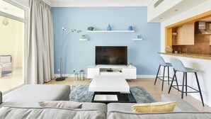A modern living room with a flat-screen TV, a white coffee table, and a sofa. There are two bar stools and a kitchen area with wooden cabinets. - Shoreline - Luxury Palm Jumeirah (Dubai)