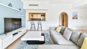 A modern living room with a flat-screen TV, a white coffee table, a grey sofa with yellow cushions, and a kitchen area with a wooden countertop and two bar stools. - Shoreline - Luxury Palm Jumeirah (Dubai)