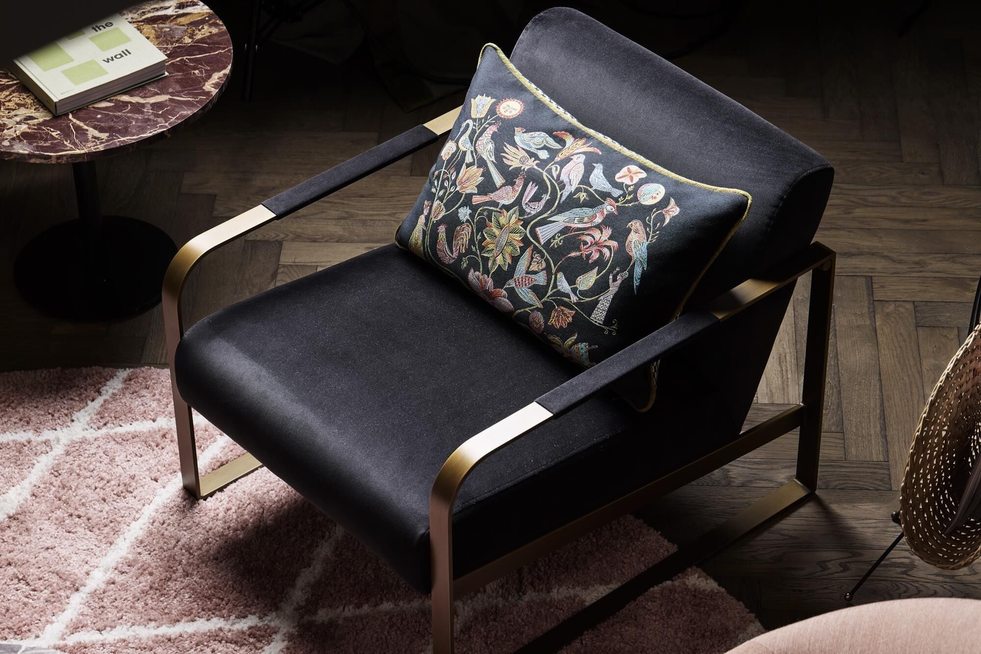 Un fauteuil noir recouvert de tissu, agrémenté d’un coussin à motifs et de bras dorés, posé sur un plancher en bois sur lequel se trouve un tapis rose.