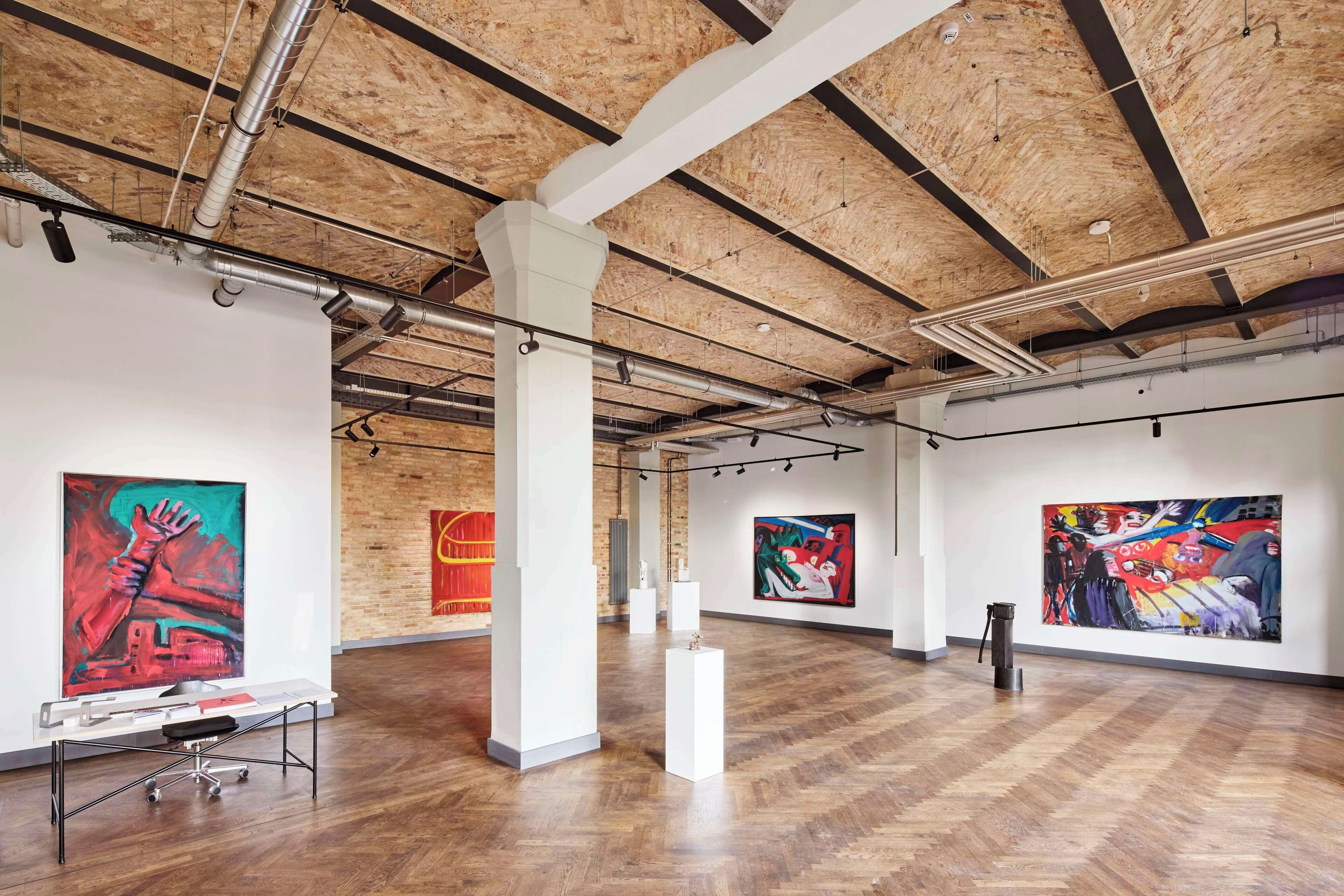 Una galería de arte con techos de madera y paredes blancas, mostrando varias pinturas de gran formato.