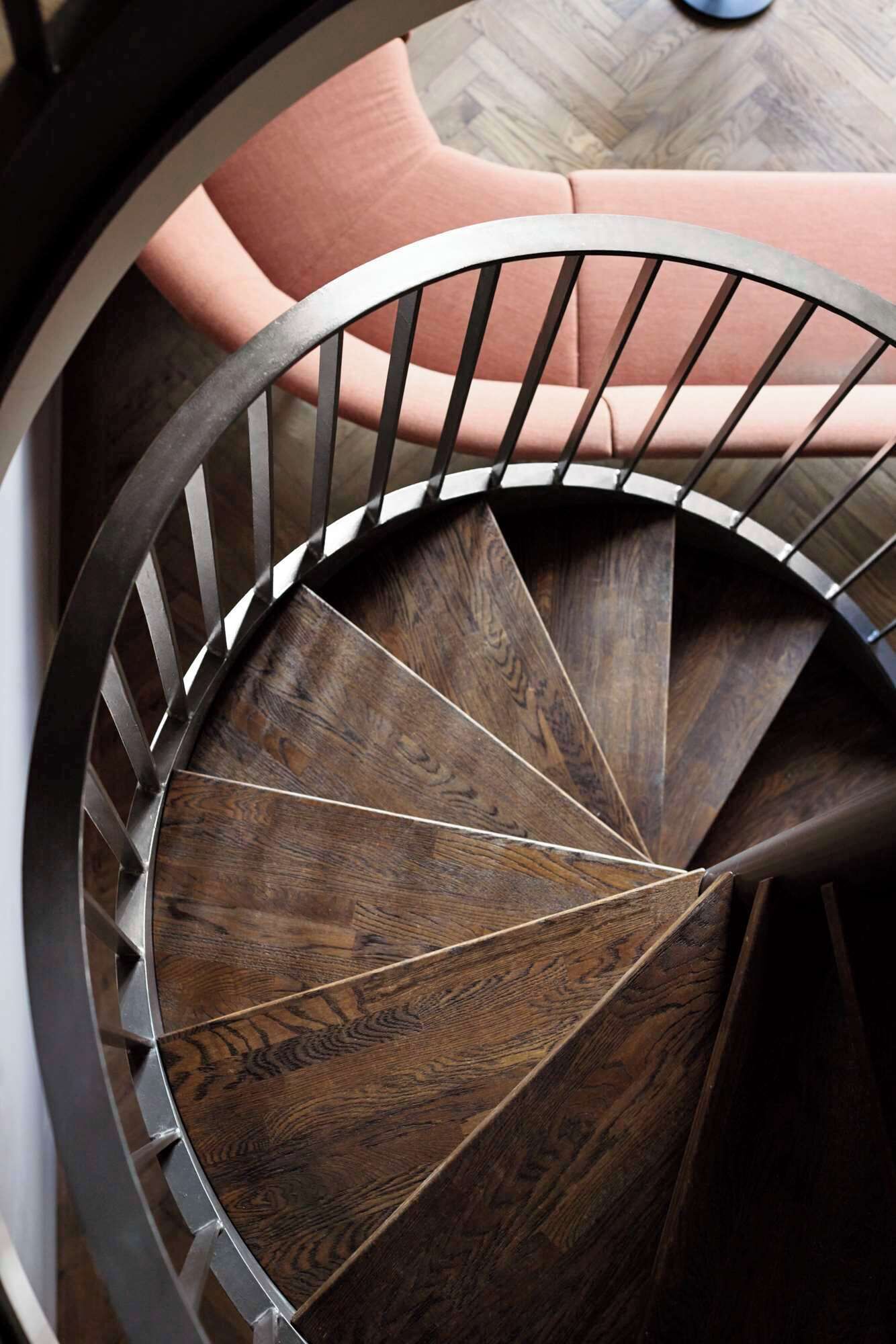Una escalera de caracol con detalles de madera y barandilla de metal, que conduce a una sala con zona de estar tapizada en tonos rosa.