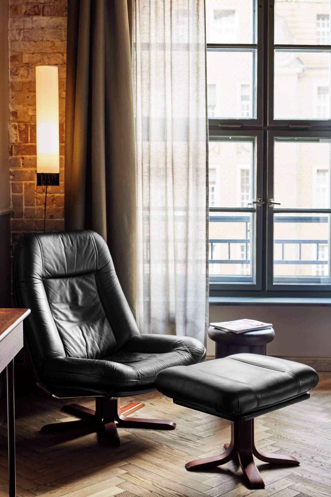 Una habitación moderna con una butaca y reposapiés de cuero, pared de ladrillo, ventana grande con cortinas y una lámpara fijada a la pared.