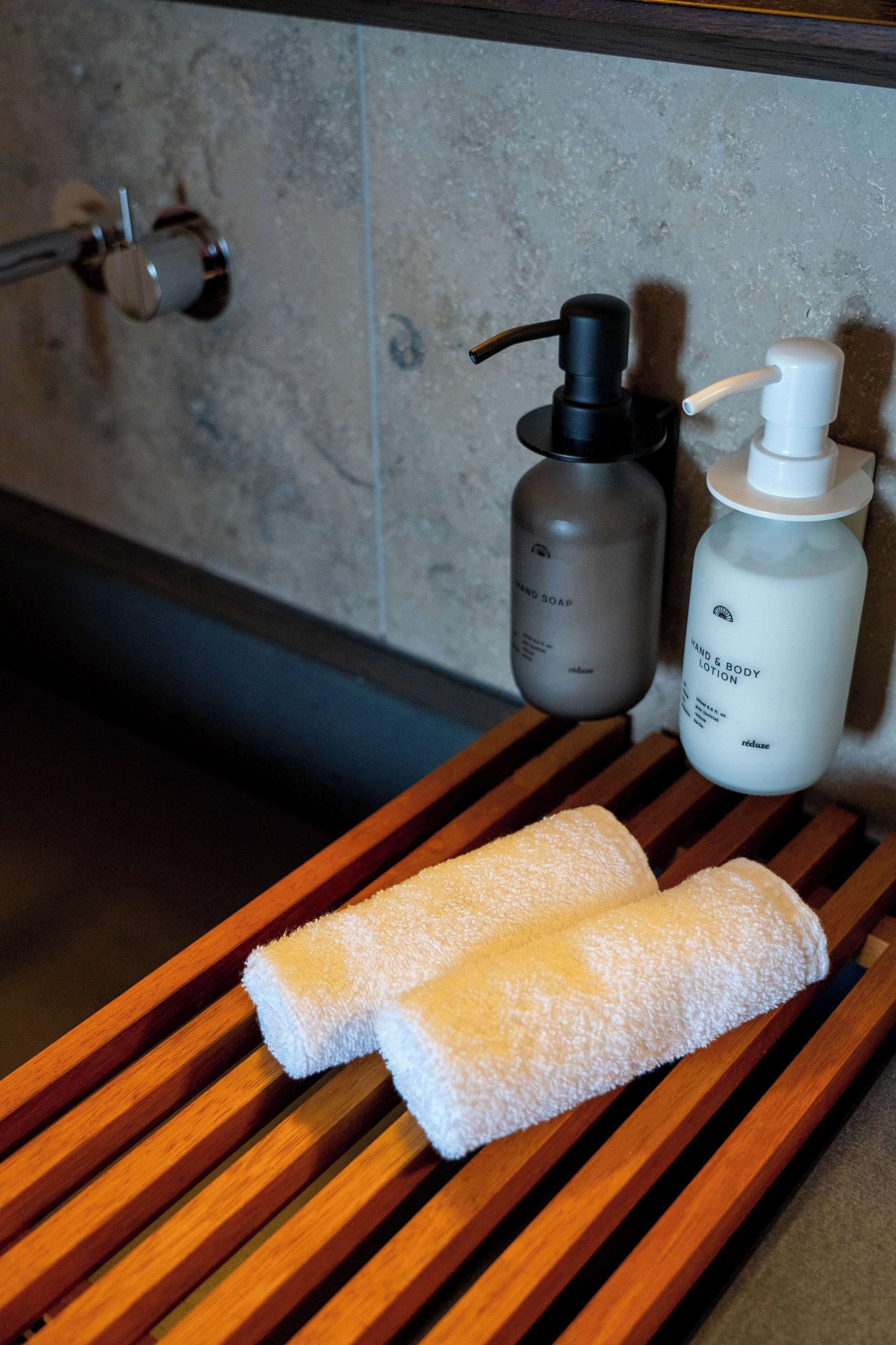 A wooden tray with two white towels, a black pump bottle, and a white bottle of lotion on a bathroom counter.