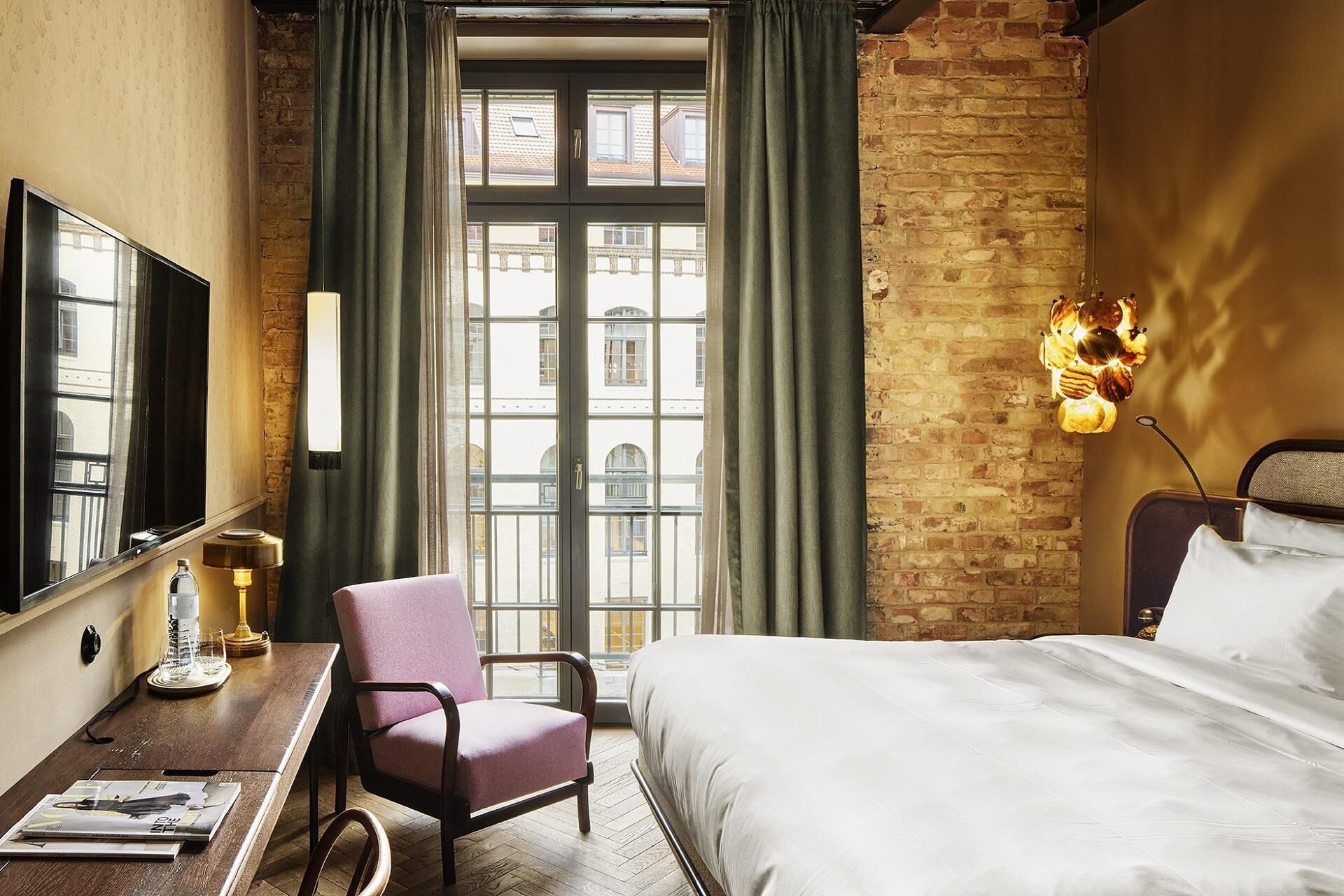 Camera d'albergo con un letto grande, una scrivania con una lampada, una sedia e vista sui palazzi attraverso una finestra con tende.