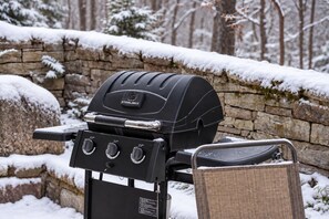 A black grill with a lid and control knobs, positioned against a stone wall with snow on top.