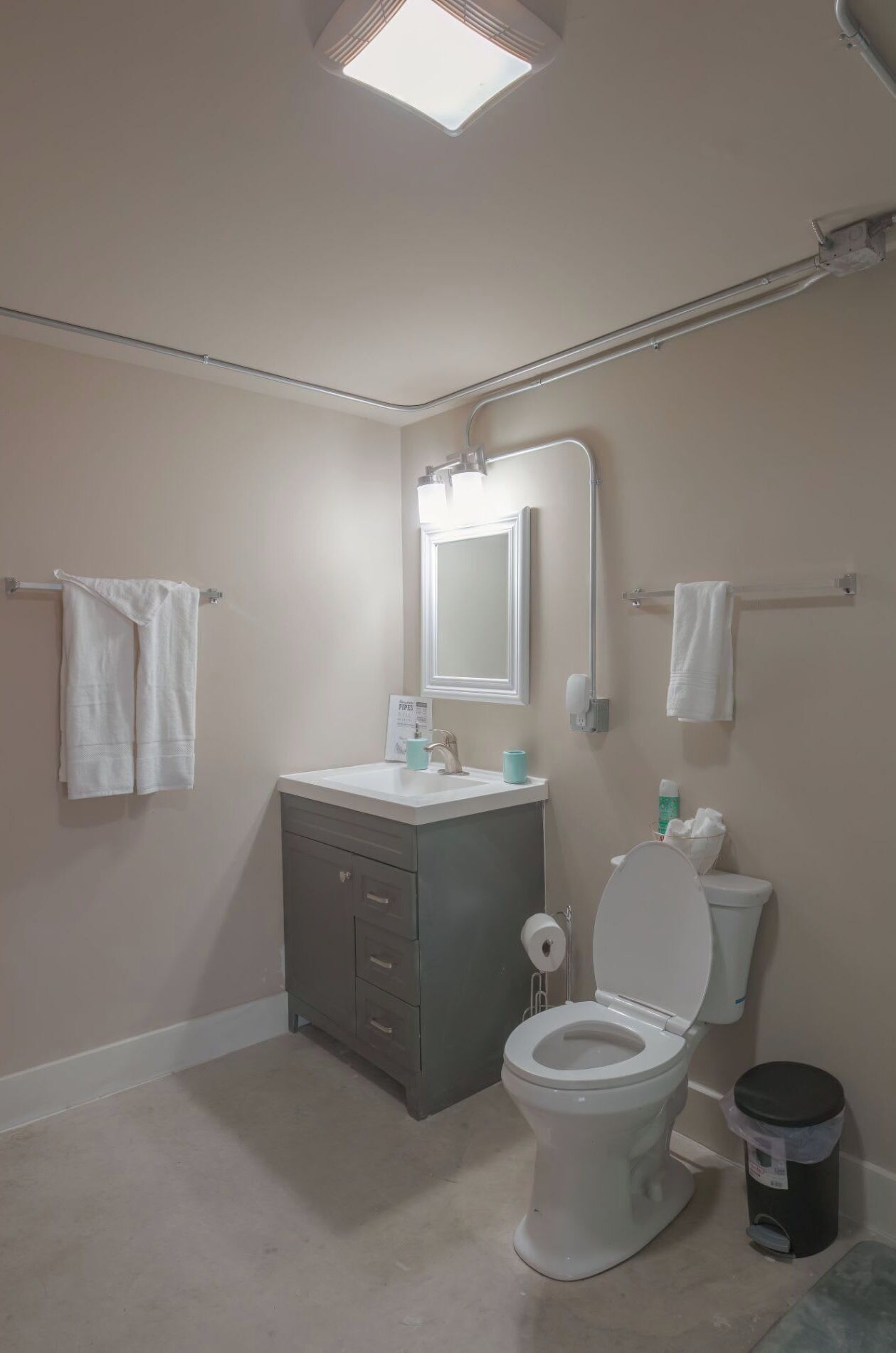 A bathroom with a toilet, sink, and mirror. There are towels hanging on the wall and a trash can.