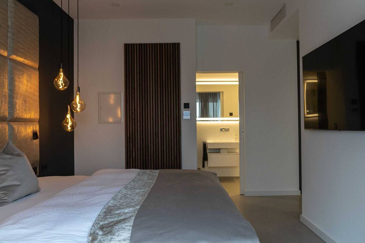 A modern hotel room with a large bed, a TV mounted on the wall, and a bathroom visible through an open door.