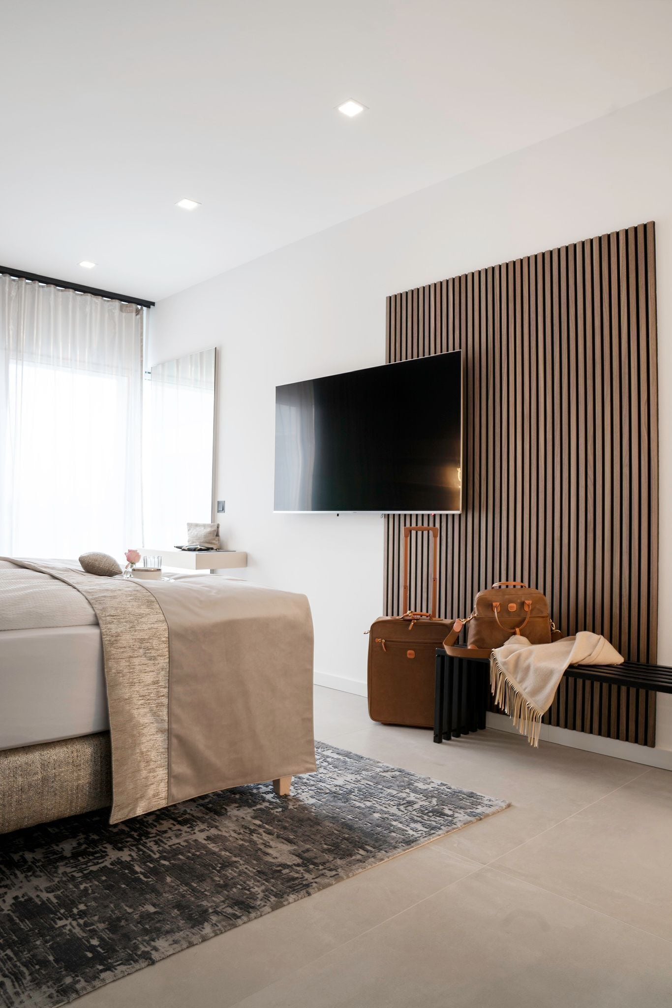 Une chambre d’hôtel moderne avec un lit, un téléviseur à écran plat fixé au mur, un décor avec des panneaux en bois, une valise et un banc.