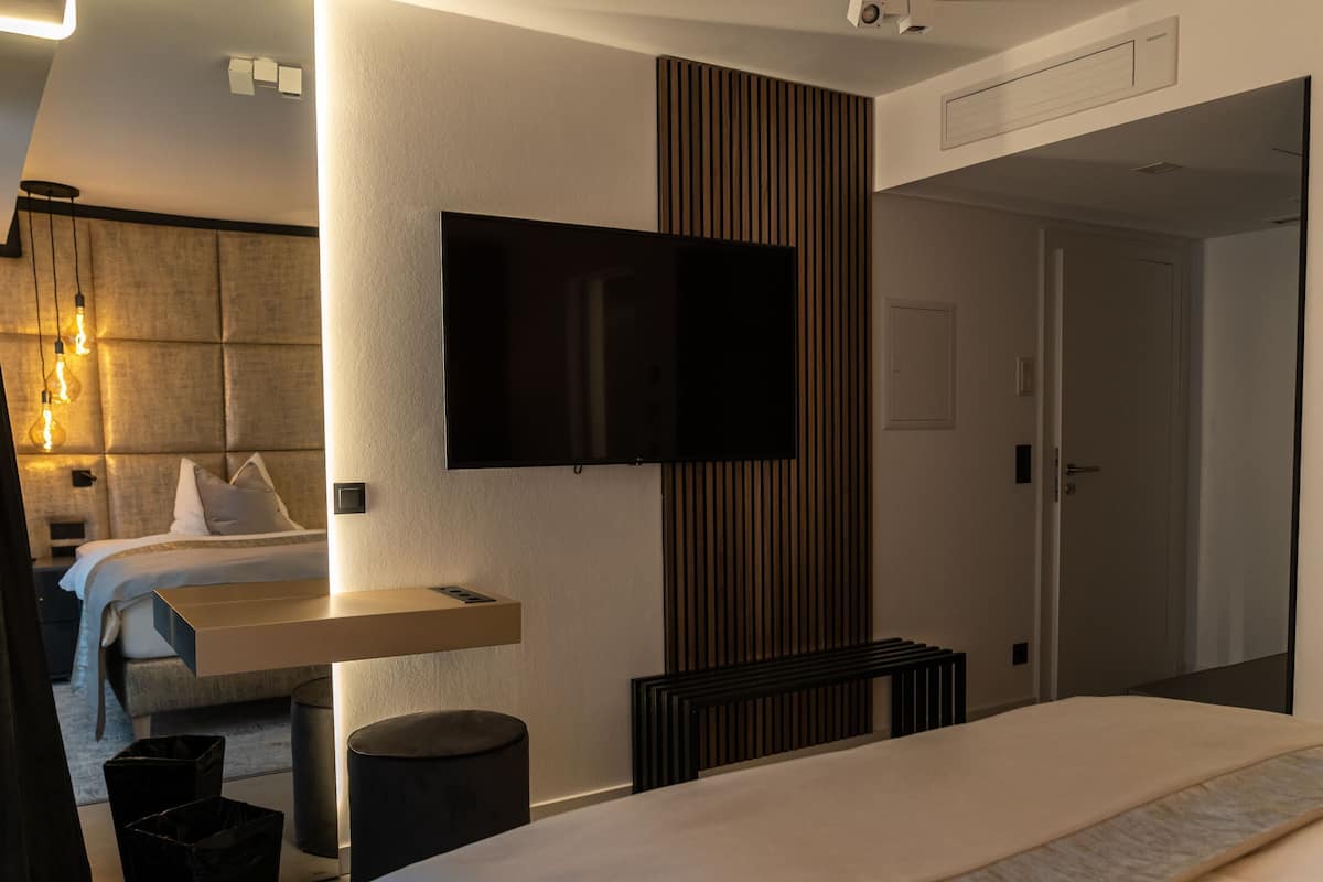 A modern hotel room with a large bed, a flat-screen TV mounted on the wall, a bedside table, a trash bin, and a door leading to another room.
