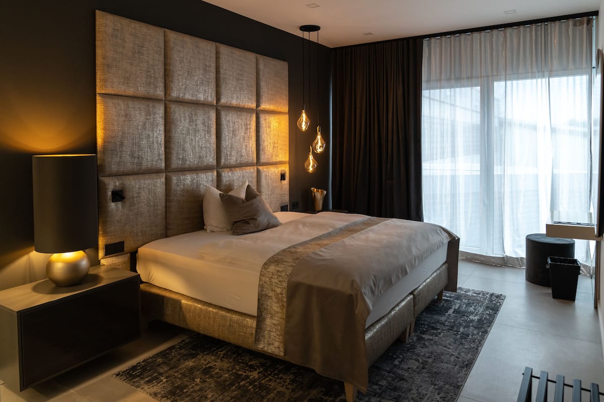 A modern hotel room with a large bed, a bedside table with a lamp, a large window with curtains, and a wall with a textured panel.