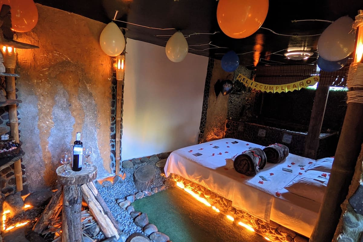 A rustic room with a stone wall, a bed with a white bedspread, a small table with a bottle and glasses, and a stone fireplace.