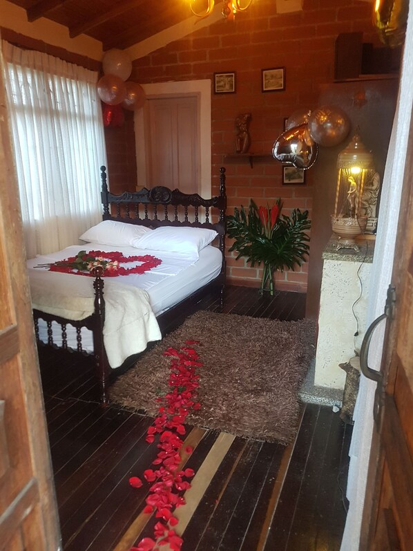 Un dormitorio con una cama, cabecera de madera, pared de ladrillo, ventana con cortinas, planta en maceta y una lámpara decorativa.
