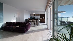 Een moderne woonkamer met een paarse bank, een eethoek en een keuken. Er is een balkon met uitzicht op het water.