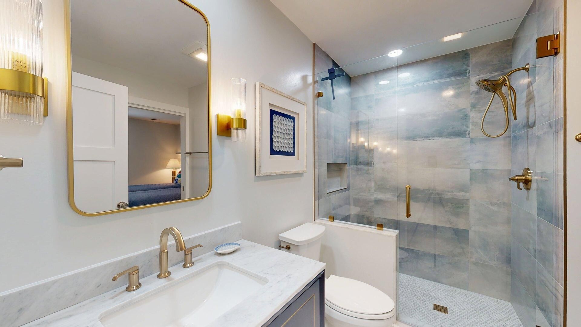 Un baño moderno con un espejo grande, un lavamanos con grifos dorados, un inodoro y una zona de ducha con cerramiento de vidrio.