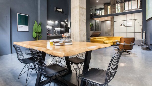 Een ruime woonkamer met een grote houten tafel, omringd door stoelen met een metalen frame, een gele bank en een houten fauteuil.