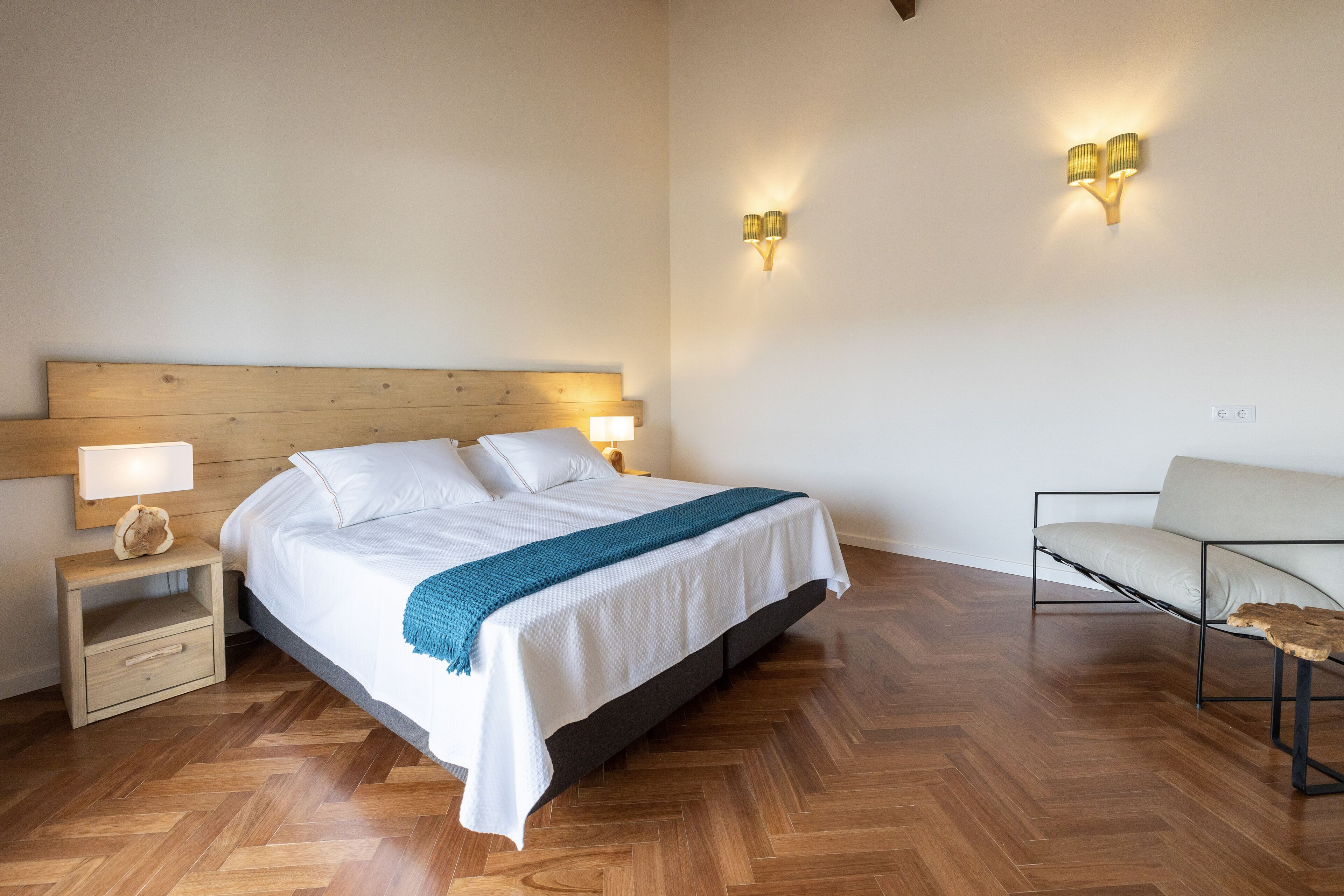 A neatly arranged bedroom with a wooden headboard bed, bedside lamps, a small wooden nightstand, and a modern chair.
