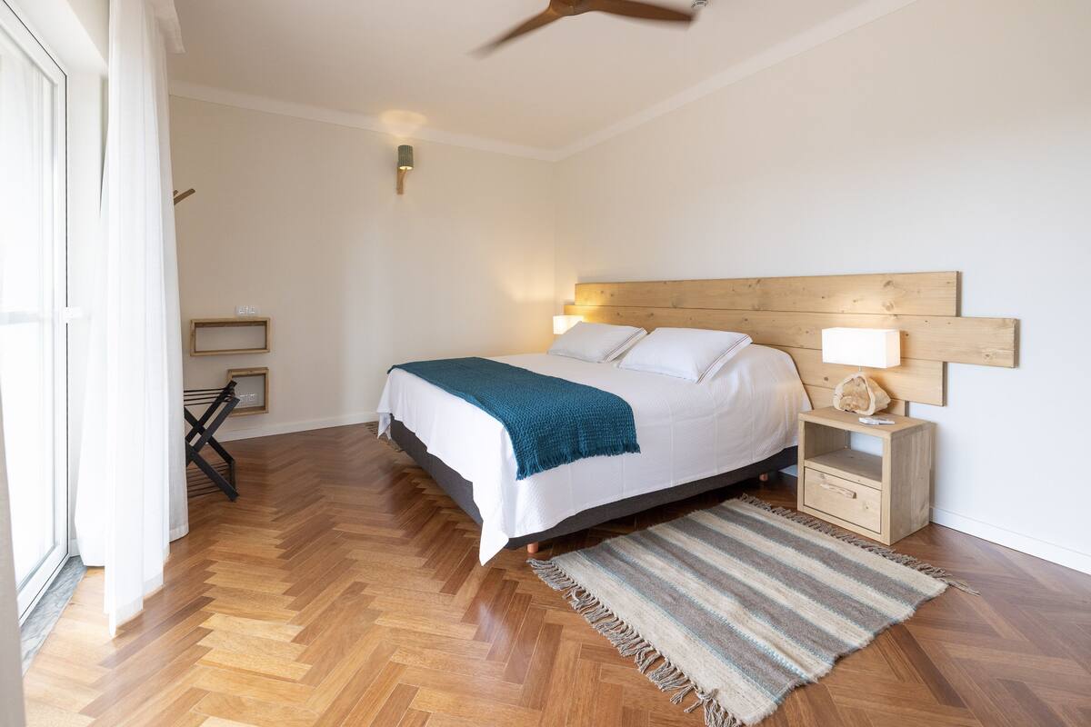 A bedroom with a wooden headboard, a bed with white bedding and a teal throw, a bedside table with a lamp, and a herringbone wooden floor.