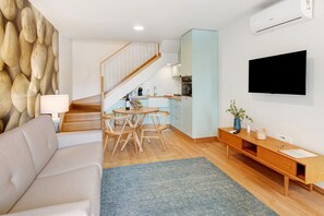 A modern living room with a sofa, a dining table with chairs, a staircase, a kitchen area, and a mounted TV.