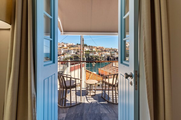 An open door to a balcony with a view of a coastal town, a pool, and outdoor furniture.