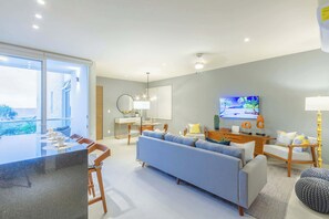 A modern living room with a sofa, a dining area, and a TV. - Ocean-View Condo, 9 Pools & Turtle Beach (Akumal)