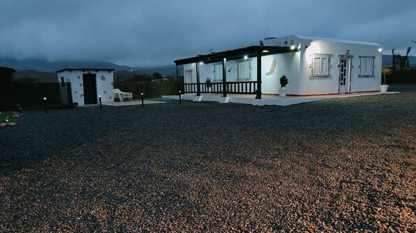 A building with a covered outdoor area, a gravel ground, and a mountainous backdrop.