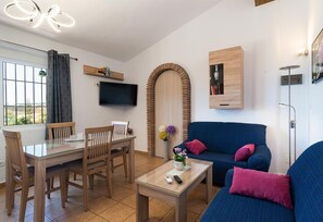 A living room with a TV, a sofa, a dining table with chairs, a small table with a vase, and a wooden cabinet.