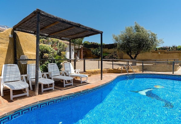 A swimming pool with a tiled edge, a covered seating area with chairs, and a clear sky.