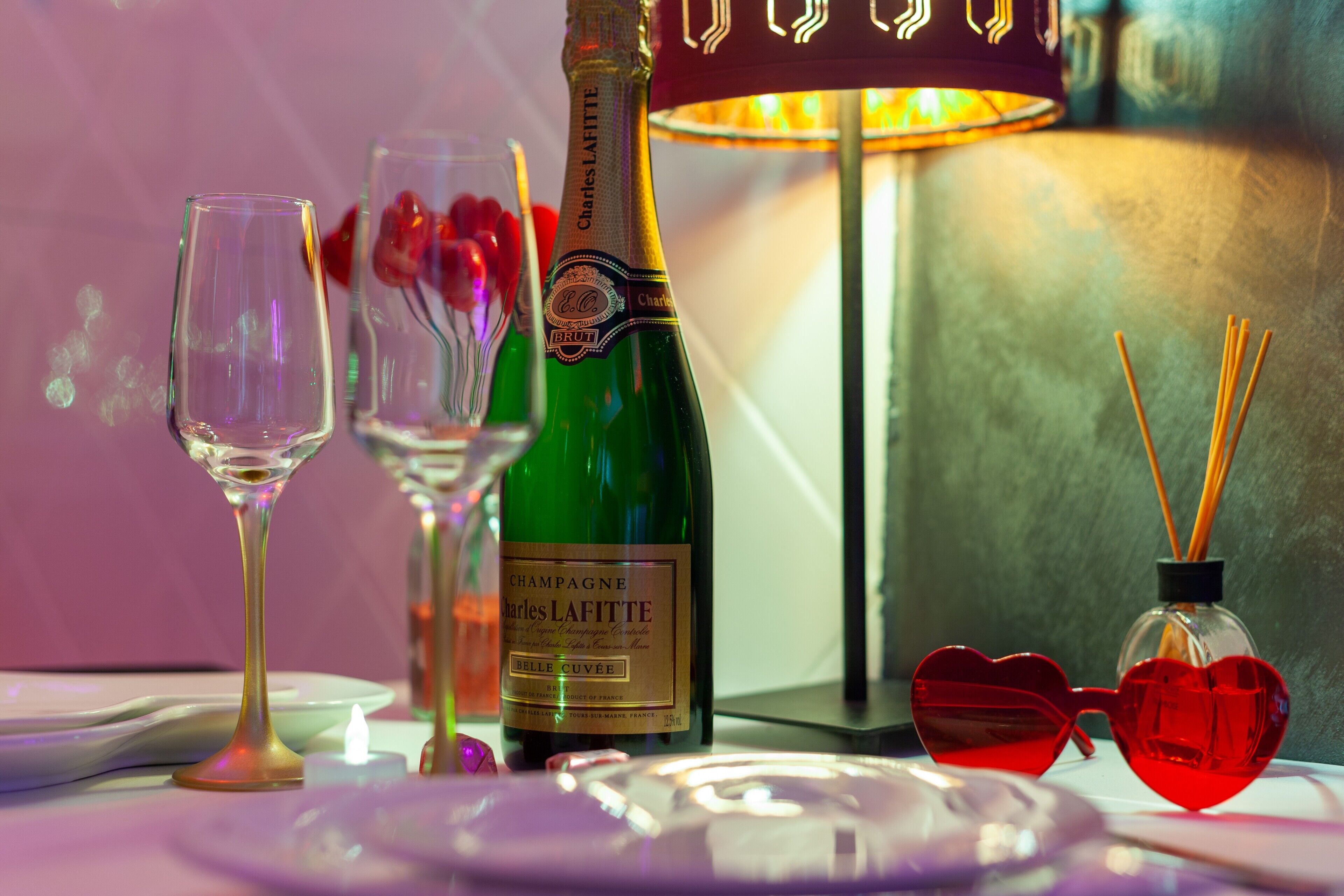 Une table dressée avec une bouteille de champagne, des verres, une paire de lunettes de soleil en forme de cœur et une bougie.
