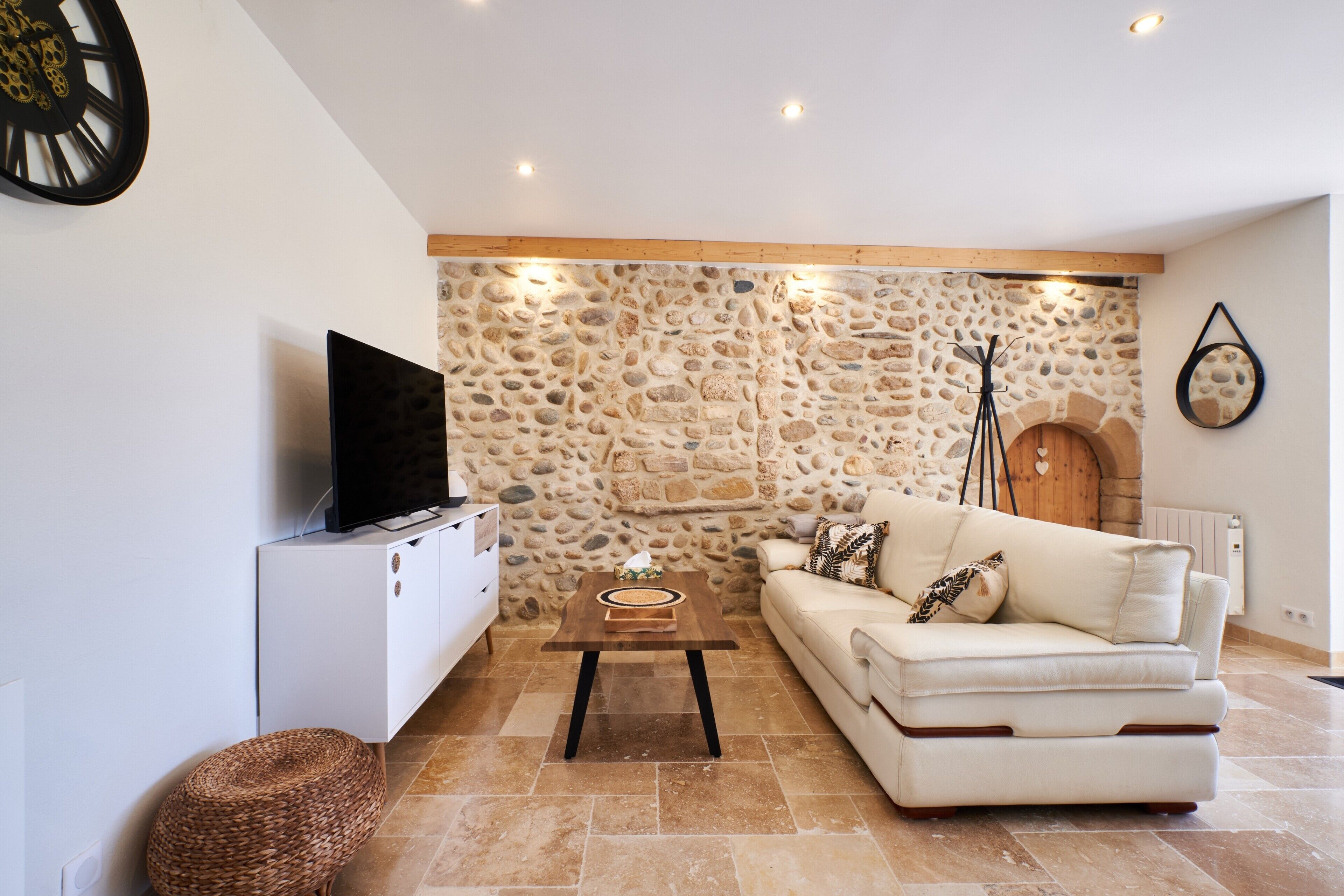 Una sala de estar con pared de piedra, un sofá blanco, un televisor sobre un mueble bajo y una lámpara de pie.